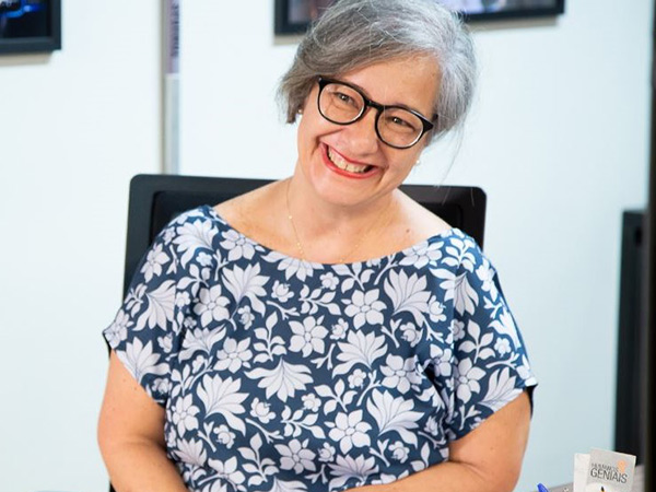 Foto de Liliane Garcez sorrindo, uma mulher de pele branca, com cabelos grisalhos e óculos de grau com armação preta